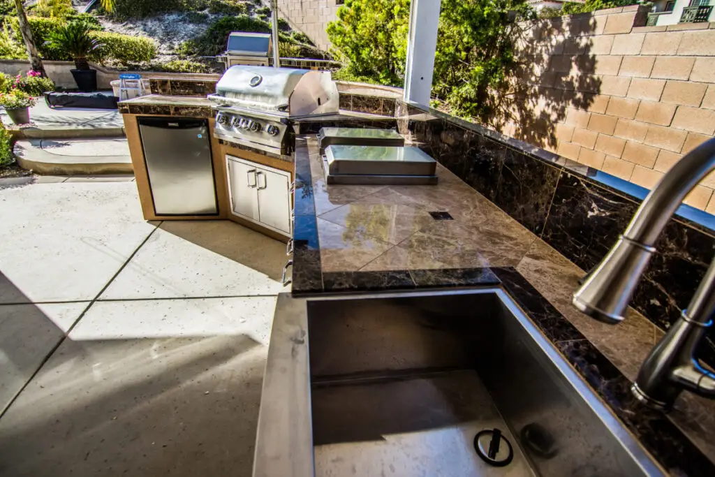 Custom outdoor kitchen workstation in Orange County featuring a stainless steel sink, built-in grill, mini fridge, marble countertop, and side burners