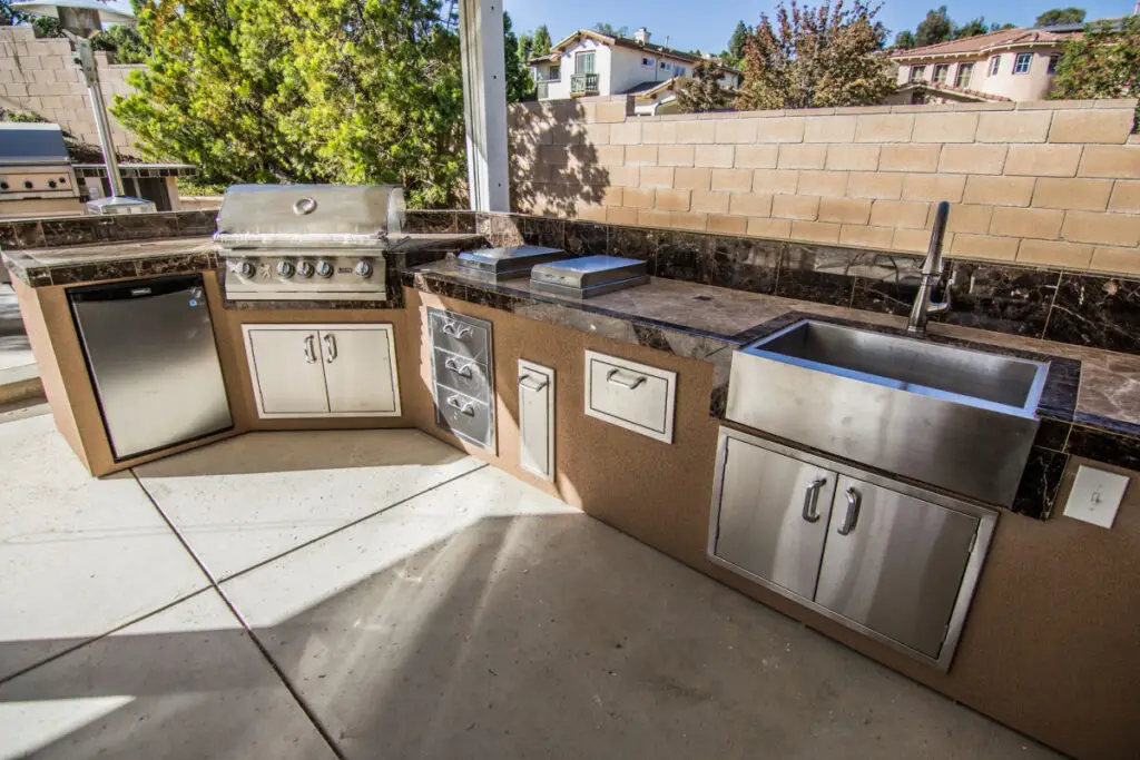 Custom-built outdoor kitchen island in Orange County featuring a grill, refrigerator, sink, marble countertops, and stainless steel storage drawers