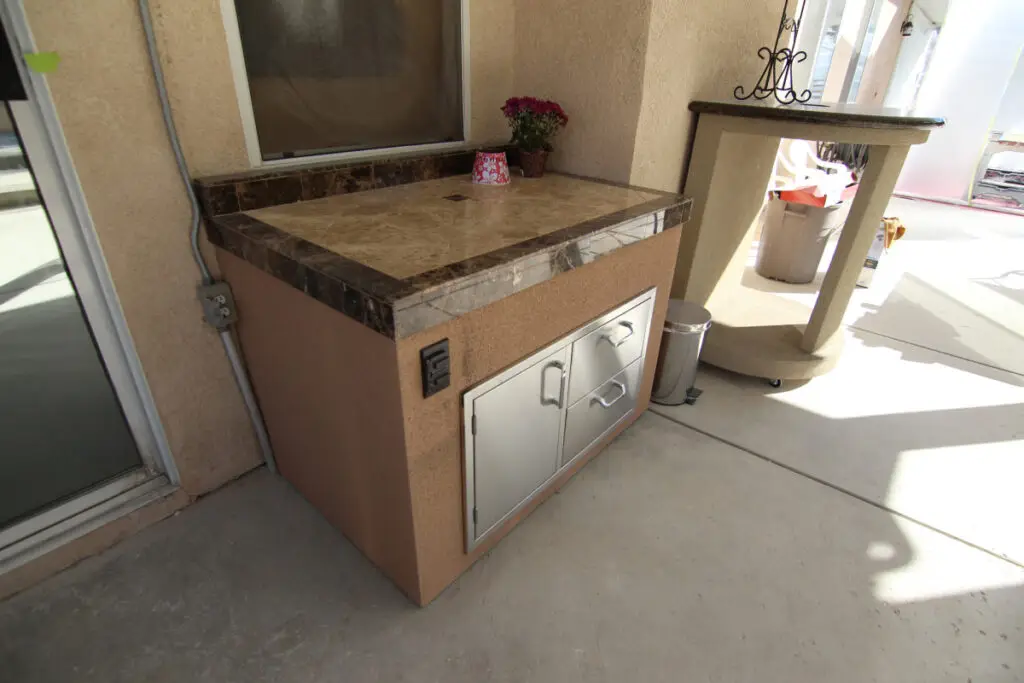 Small outdoor kitchen storage unit with marble countertop, stainless steel drawers, and electrical outlet under a covered patio in Orange County
