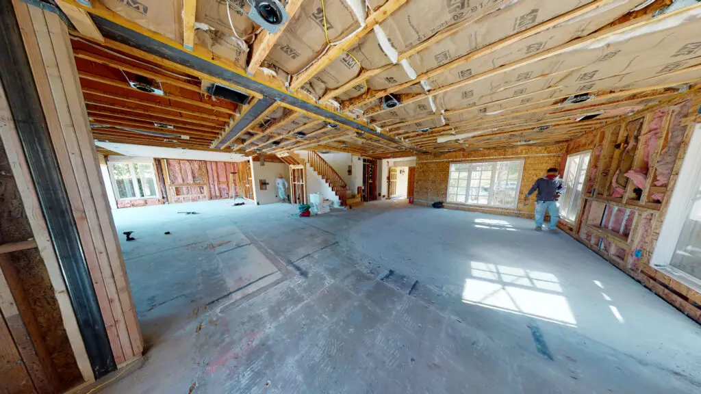 Large room addition under construction with exposed ceiling joists, insulation, framing, and workers preparing interior for drywall installation