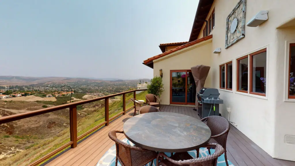 Spacious hillside deck with brown composite decking, glass railing, outdoor dining set, and built-in BBQ grill overlooking panoramic valley views in Orange County