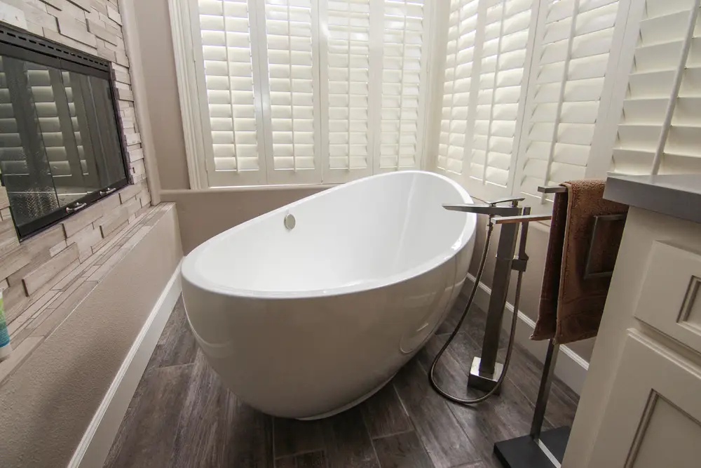 Elegant bathroom remodel featuring a freestanding white soaking tub beside a modern stone fireplace and surrounded by plantation shutters for natural light and privacy