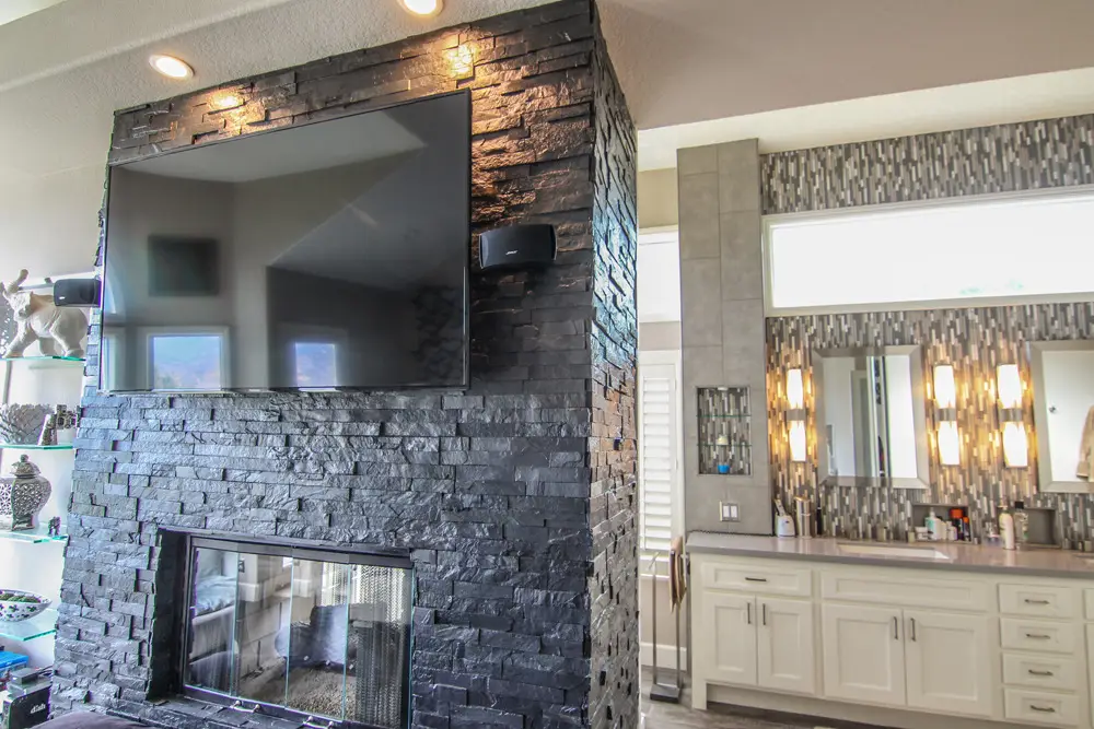Modern luxury bathroom remodel featuring a slate tile double-sided fireplace with mounted TV, dual sink vanity with white cabinets, mosaic tile backsplash, and ambient wall lighting