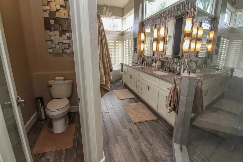 Spacious modern bathroom remodel featuring a long double-sink vanity with white cabinets, tile backsplash, warm lighting, wood-look tile flooring, and separate toilet room
