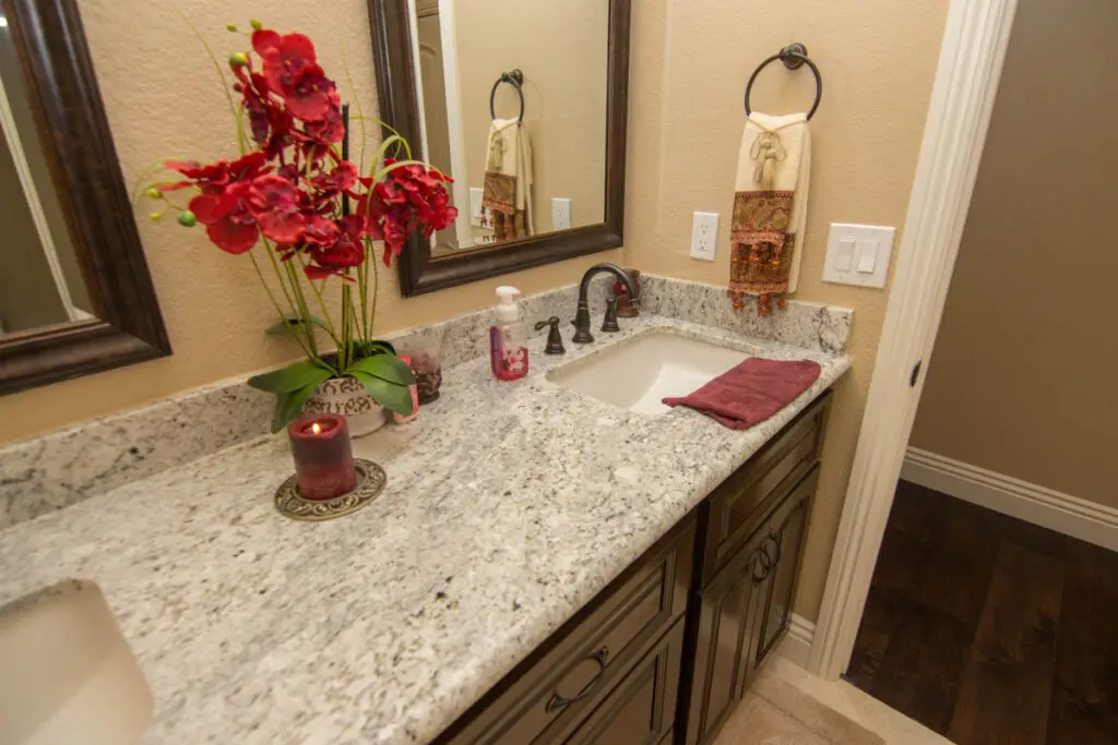 Close-up of a remodeled bathroom vanity with granite countertop, oil-rubbed bronze faucet, red floral arrangement, and decorative hand towels