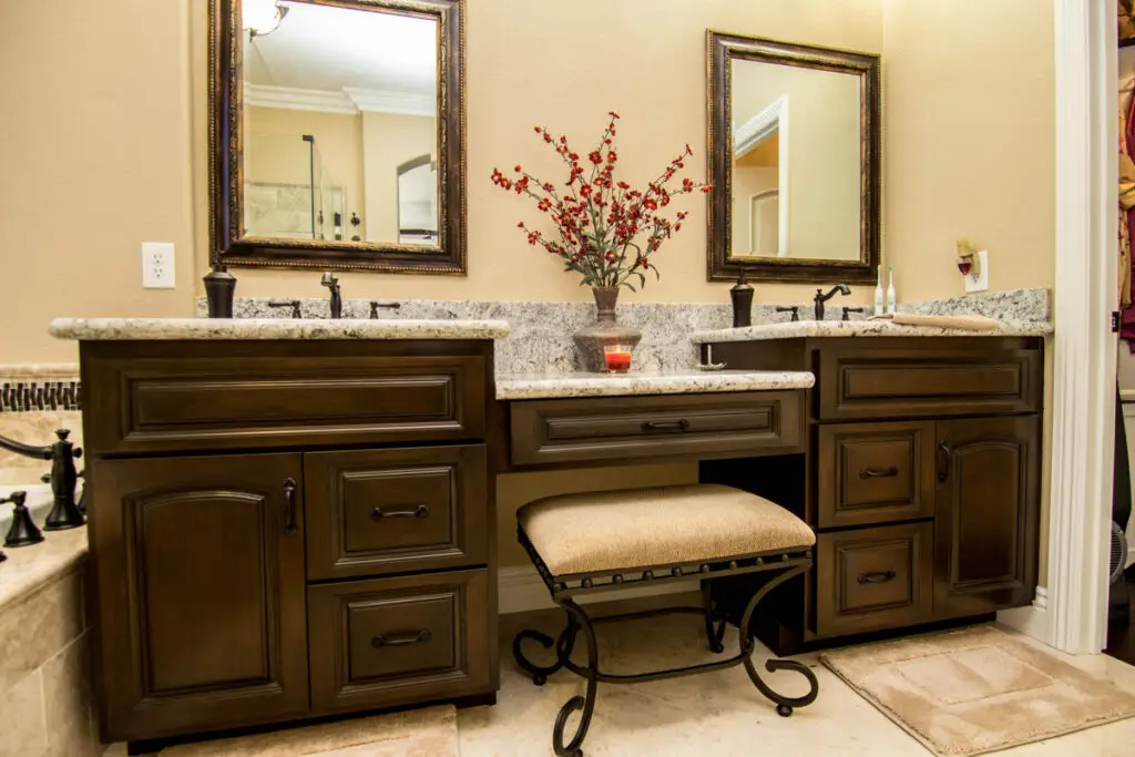 Refined bathroom vanity remodel featuring dark wood cabinetry, granite counters, framed dual mirrors, and a built-in makeup station with a cushioned stool