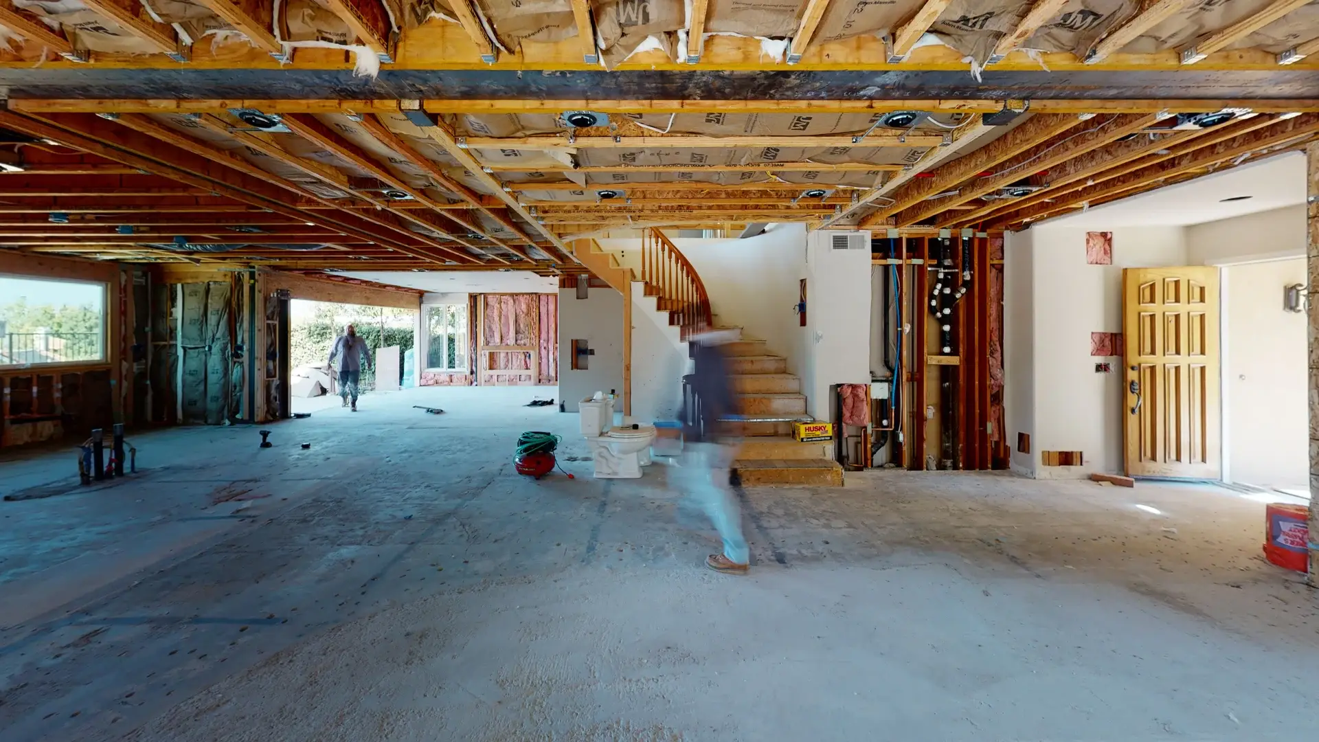 Full interior view of a whole house makeover showing exposed ceiling beams, insulation installation, rough plumbing, and staircase framing with workers on site.