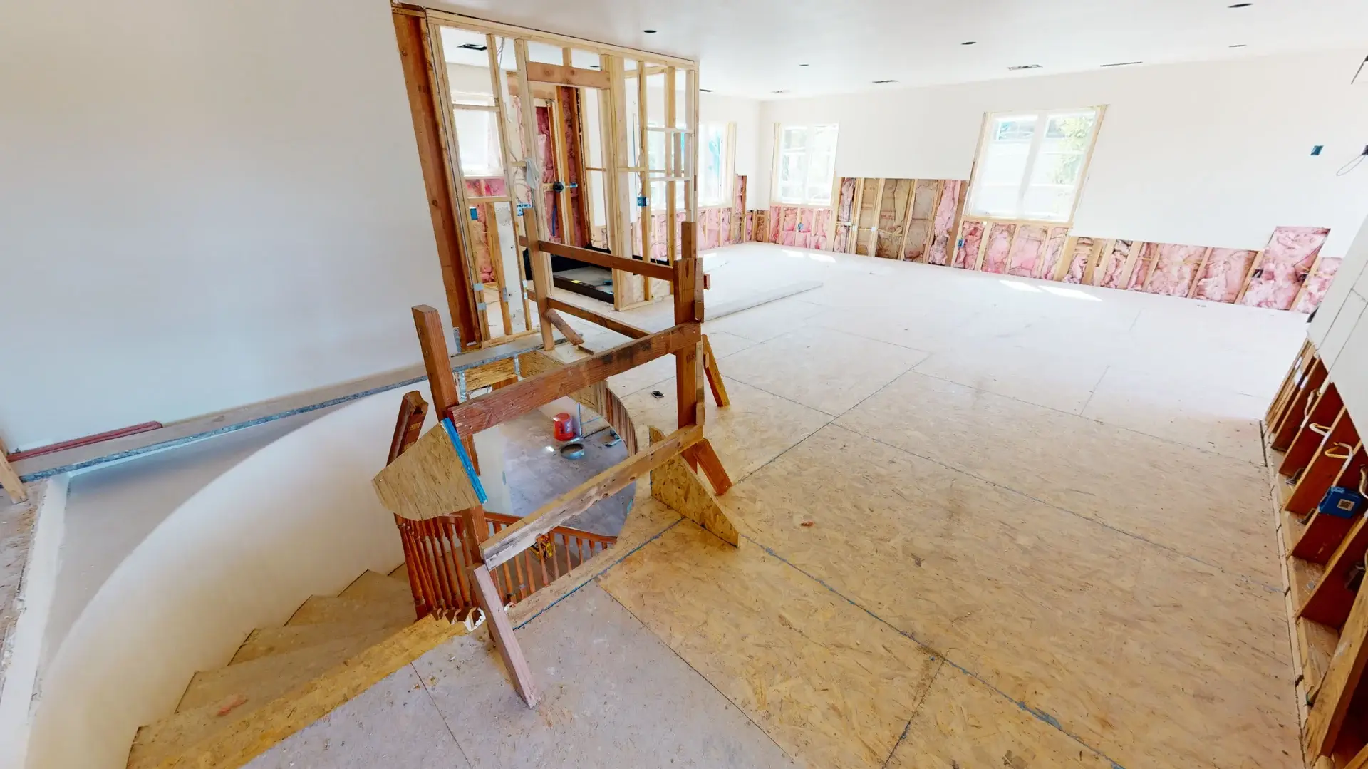 Construction progress of a whole house makeover showing plywood subflooring, partial wall framing, exposed insulation, and curved staircase access on the second floor, part of a full home remodel project in Orange County, CA.