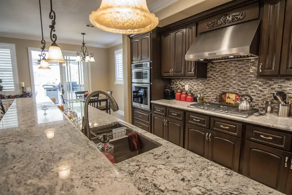 Spacious remodeled kitchen featuring an oversized granite island with an undermount sink, dark wood cabinetry, stainless steel appliances, mosaic tile backsplash, and pendant lighting—designed for modern cooking and entertaining.