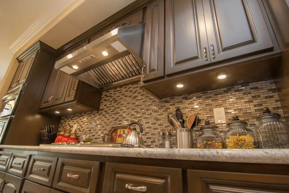 Close-up view of a luxury kitchen remodel featuring custom dark wood cabinets, stainless steel range hood, under-cabinet lighting, and a mosaic tile backsplash with marble countertop—showcasing premium finishes and elegant design.