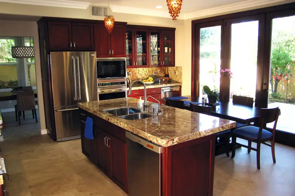 Modern kitchen remodel featuring rich cherry wood cabinets, stainless steel refrigerator and appliances, granite countertop island with a double sink, and floor-to-ceiling glass doors for natural light and indoor-outdoor living.