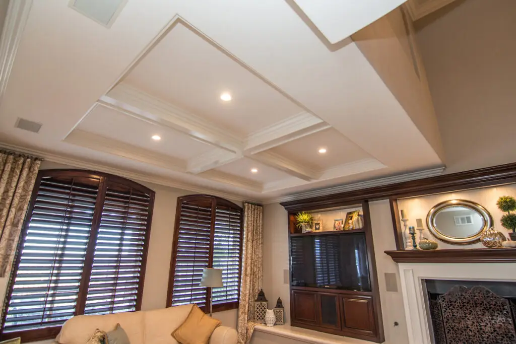 View from below of a luxurious room addition featuring a coffered ceiling, dark wood built-in entertainment center, fireplace with mirror, and large arched windows with plantation shutters.