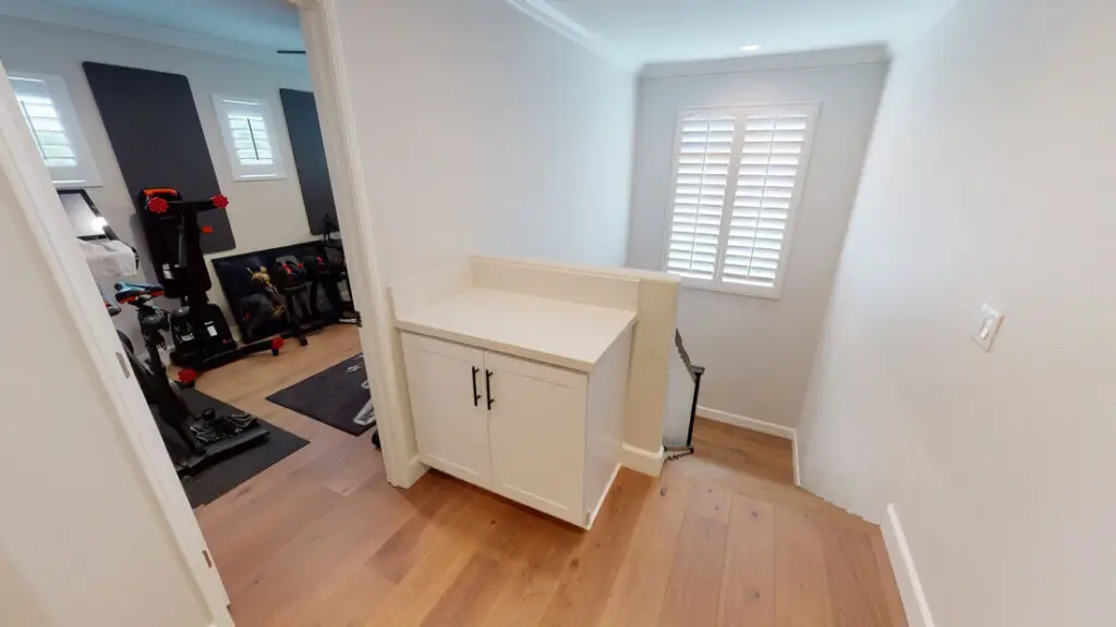Bright hallway view leading to a custom-built home gym addition with hardwood floors, white shaker cabinet, and plantation shutters.