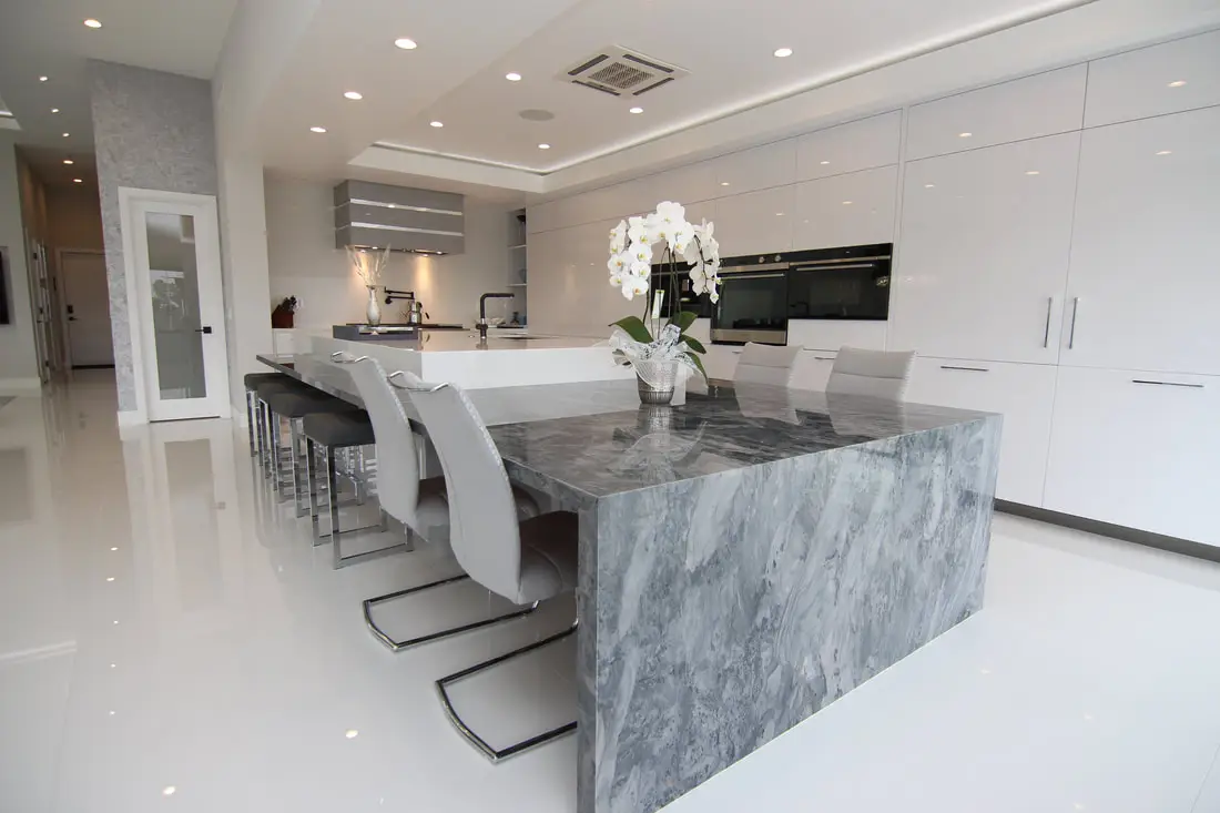 Contemporary kitchen featuring a gray marble waterfall island with seating, high-gloss white cabinetry, built-in ovens, and sleek recessed lighting