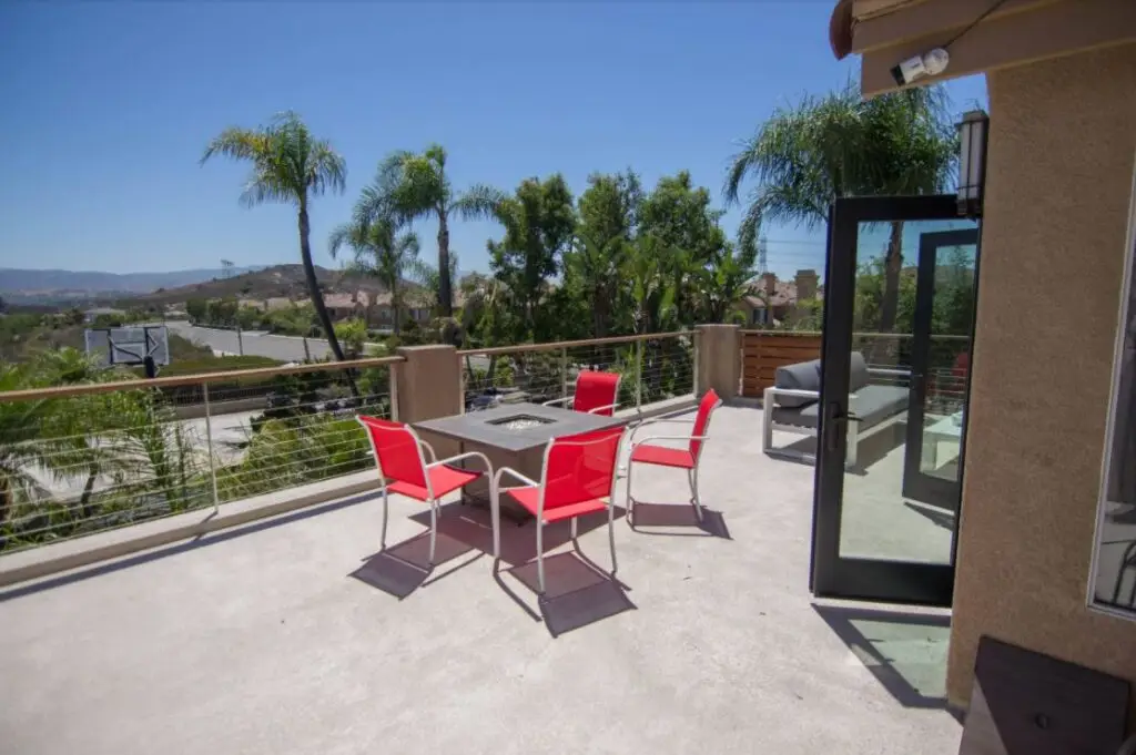 Spacious rooftop patio with red modern chairs around a fire pit table, glass doors open to seating area, overlooking lush landscaping and hills in the background