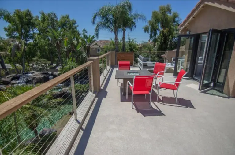 Contemporary rooftop deck featuring a sleek fire pit table, bold red patio chairs, cable railing, and views of lush landscaping with a waterfall below