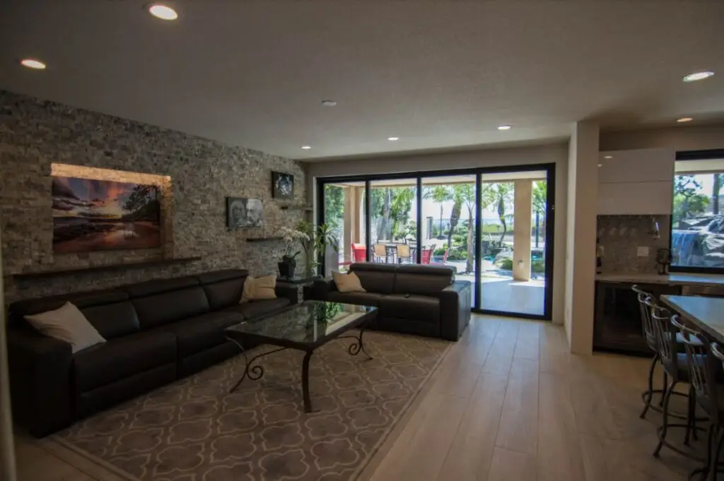Spacious California room interior with leather sectionals, stone feature wall, and wide sliding glass doors opening to a luxury patio with poolside seating and tropical landscaping