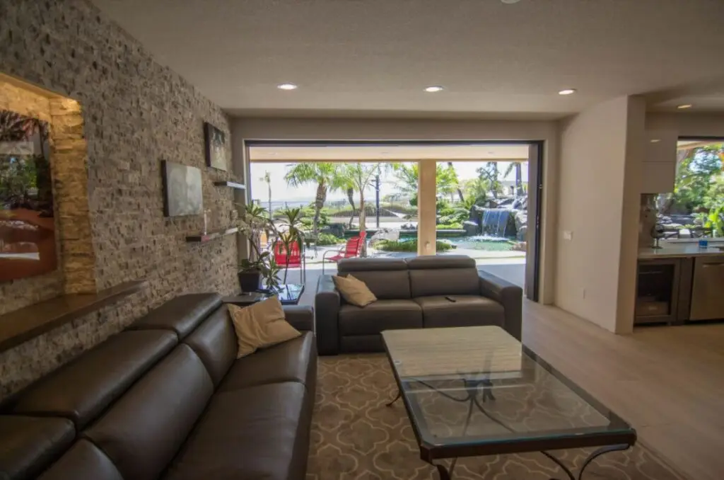 Interior view of a modern California room featuring large sliding glass doors, leather sofas, stone accent wall, and a clear view of the backyard pool with waterfall and patio seating